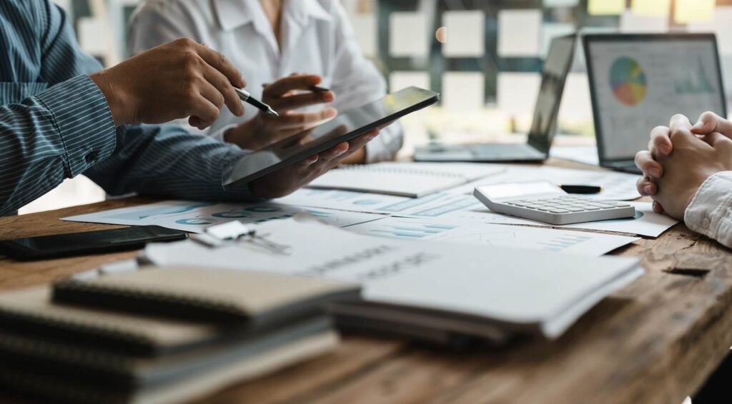 Close-up of a business meeting with people discussing financial documents, using a tablet, calculator, and laptops at a wooden table.