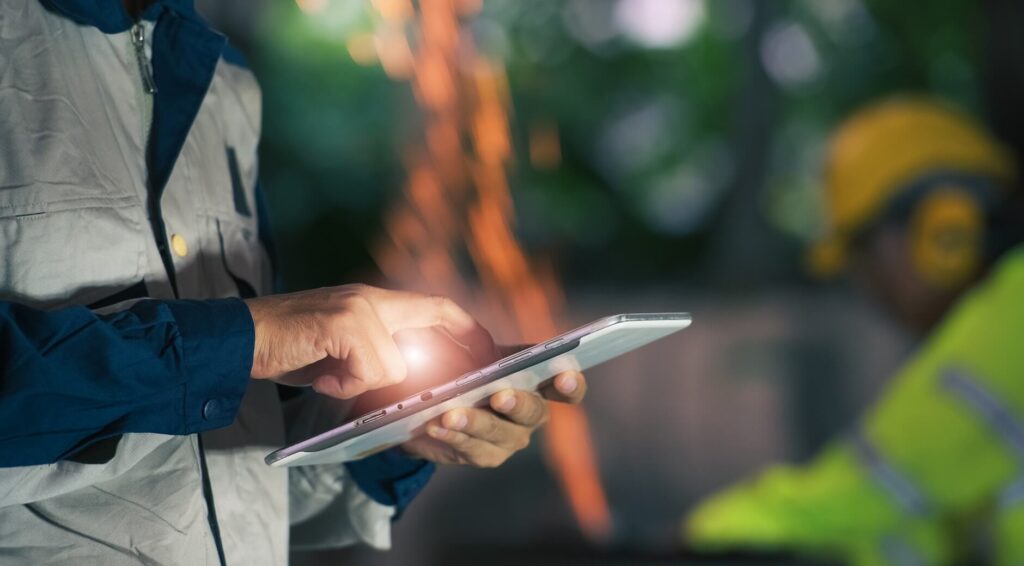 Engineer using a tablet at an industrial site with a worker and welding sparks in the background.